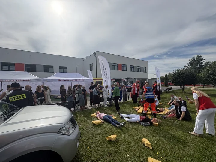 Pokaz ratownictwa medycznego na trawie przed nowoczesnym budynkiem szpitala, z udziałem służb ratunkowych, symulowanymi poszkodowanymi i grupą obserwatorów.