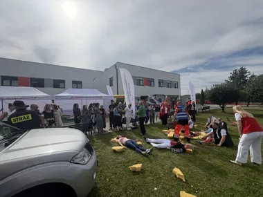 Pokaz ratownictwa medycznego na trawie przed nowoczesnym budynkiem szpitala, z udziałem służb ratunkowych, symulowanymi poszkodowanymi i grupą obserwatorów.