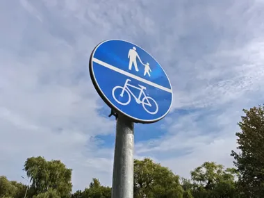 Niebieski okrągły znak drogowy wskazujący wspólną ścieżkę dla pieszych i rowerzystów, zamontowany na metalowym słupku na tle częściowo zachmurzonego nieba i drz